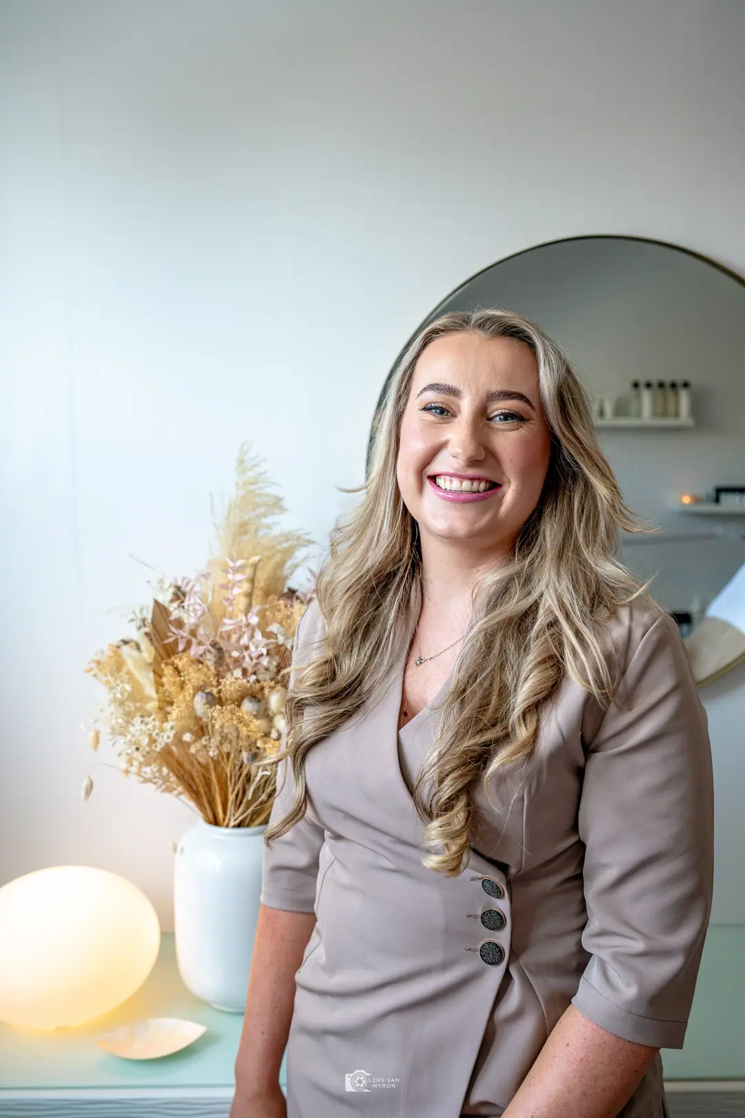 Celine lacht in haar behandelruimte, naast droogbloemen en een ronde spiegel.