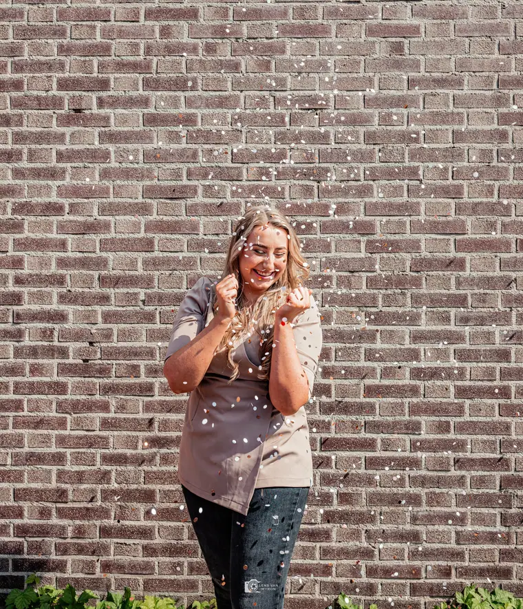 Celine lacht in de confetti voor een bakstenen muur.