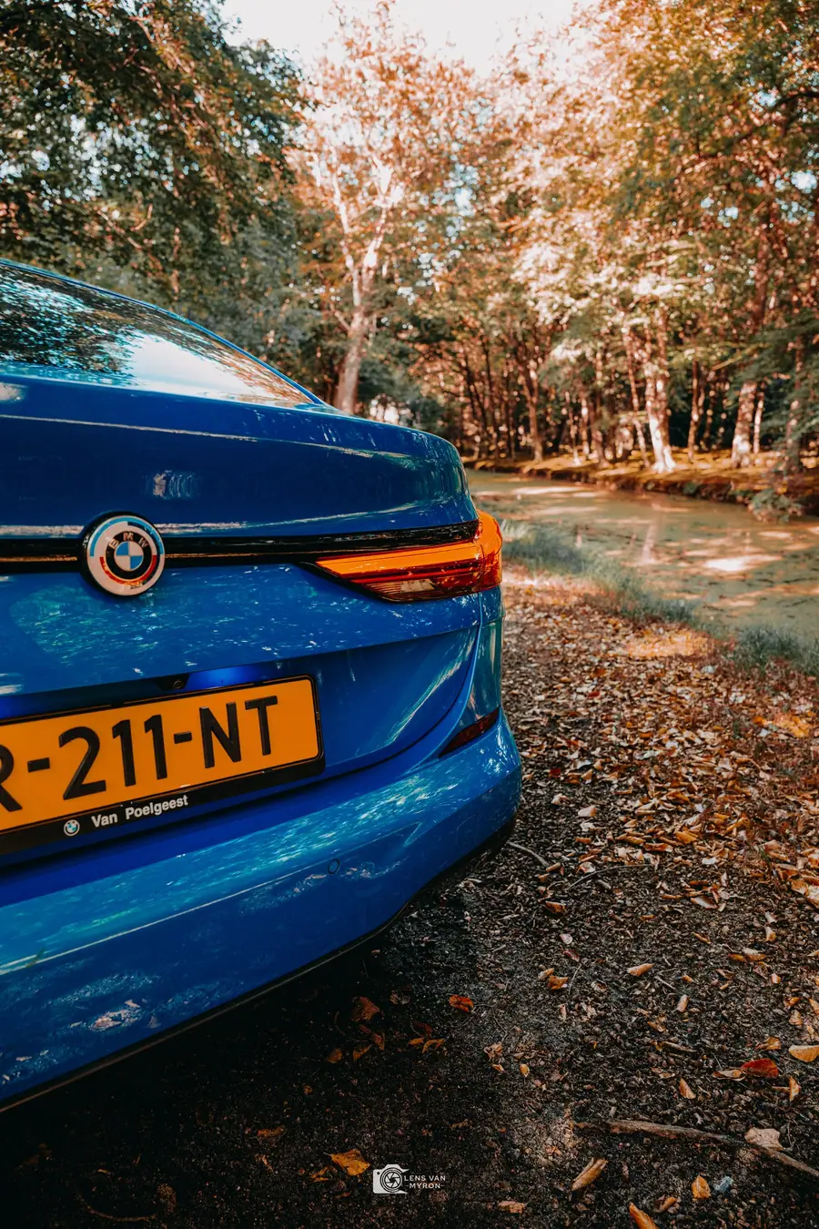 Detail van de achterkant van een blauwe BMW 2 Serie met embleem en achterlicht, met herfstbomen en een vijver in de verte
