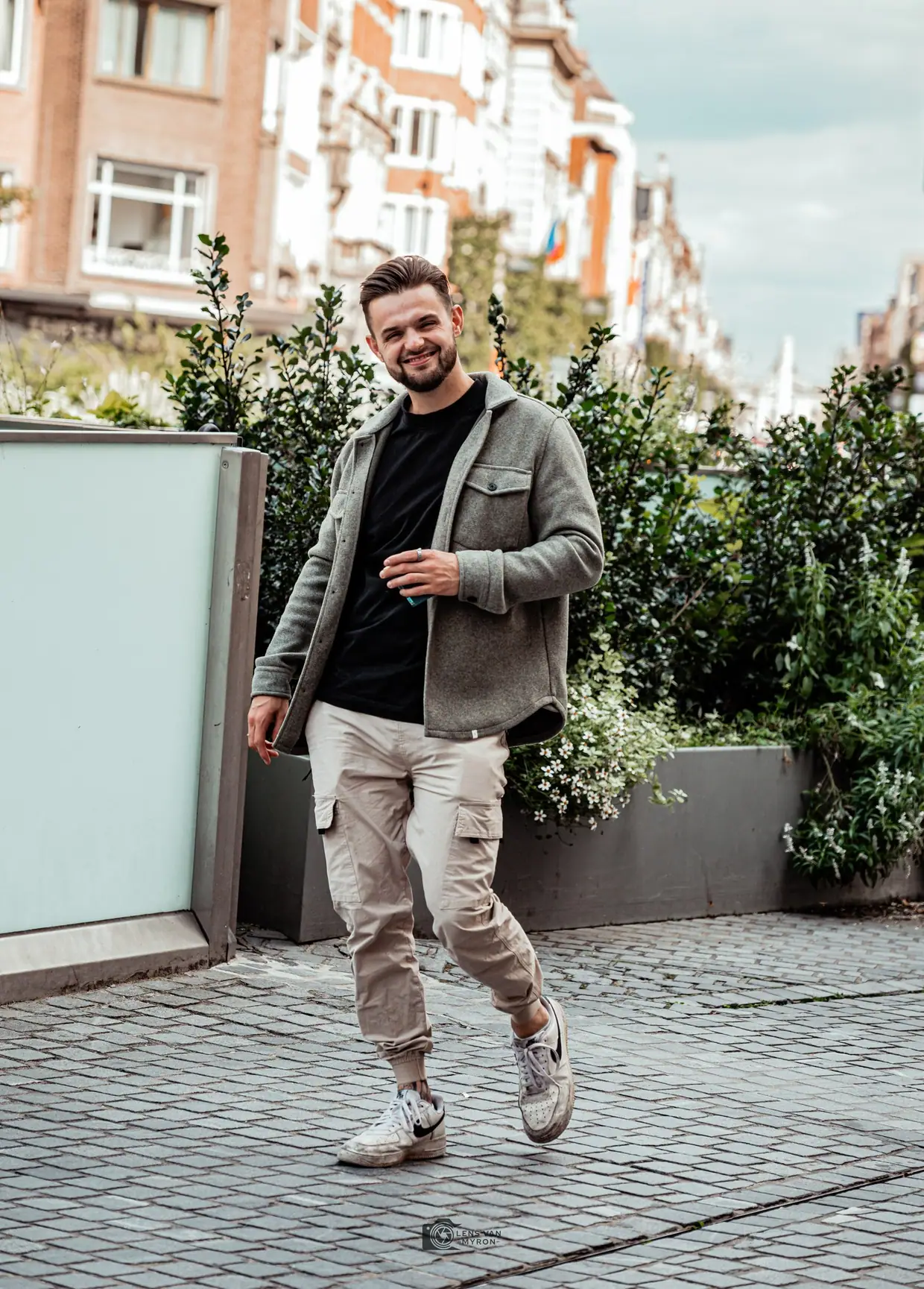 Bobby loopt lachend over een klinkerstraat langs plantenbakken, in een grijs overshirt, zwart T-shirt, beige cargobroek en witte sneakers.