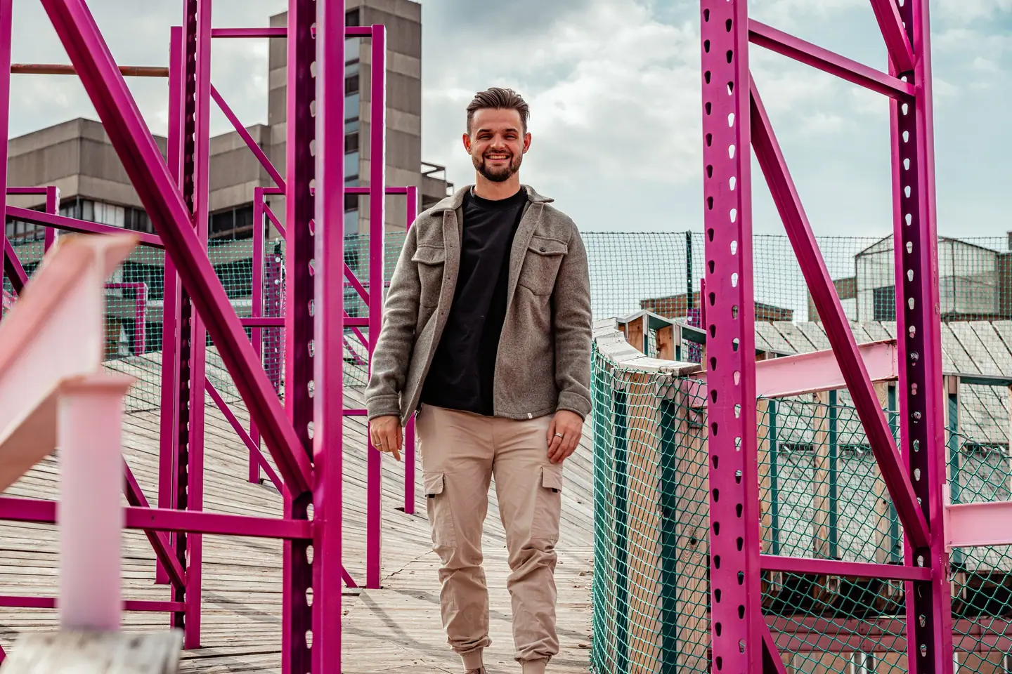 Bobby loopt glimlachend over een houten loopbrug tussen felroze stalen balken, met betonnen gebouwen en een bewolkte lucht op de achtergrond.