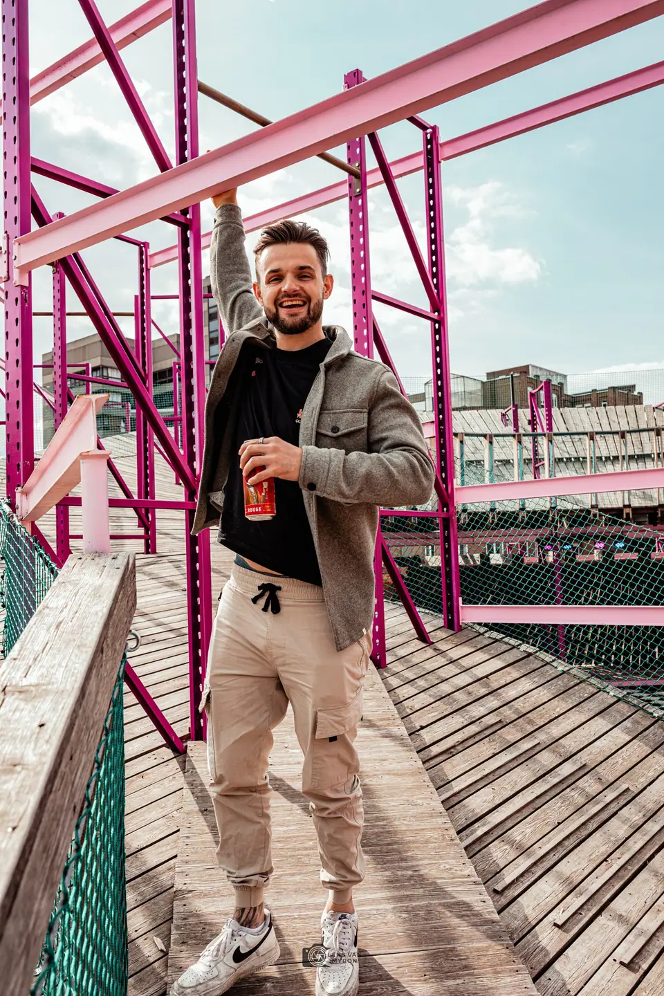 Bobby hangt lachend met één hand aan een roze stalen balk en houdt in de andere een blikje vast, op een houten loopbrug.