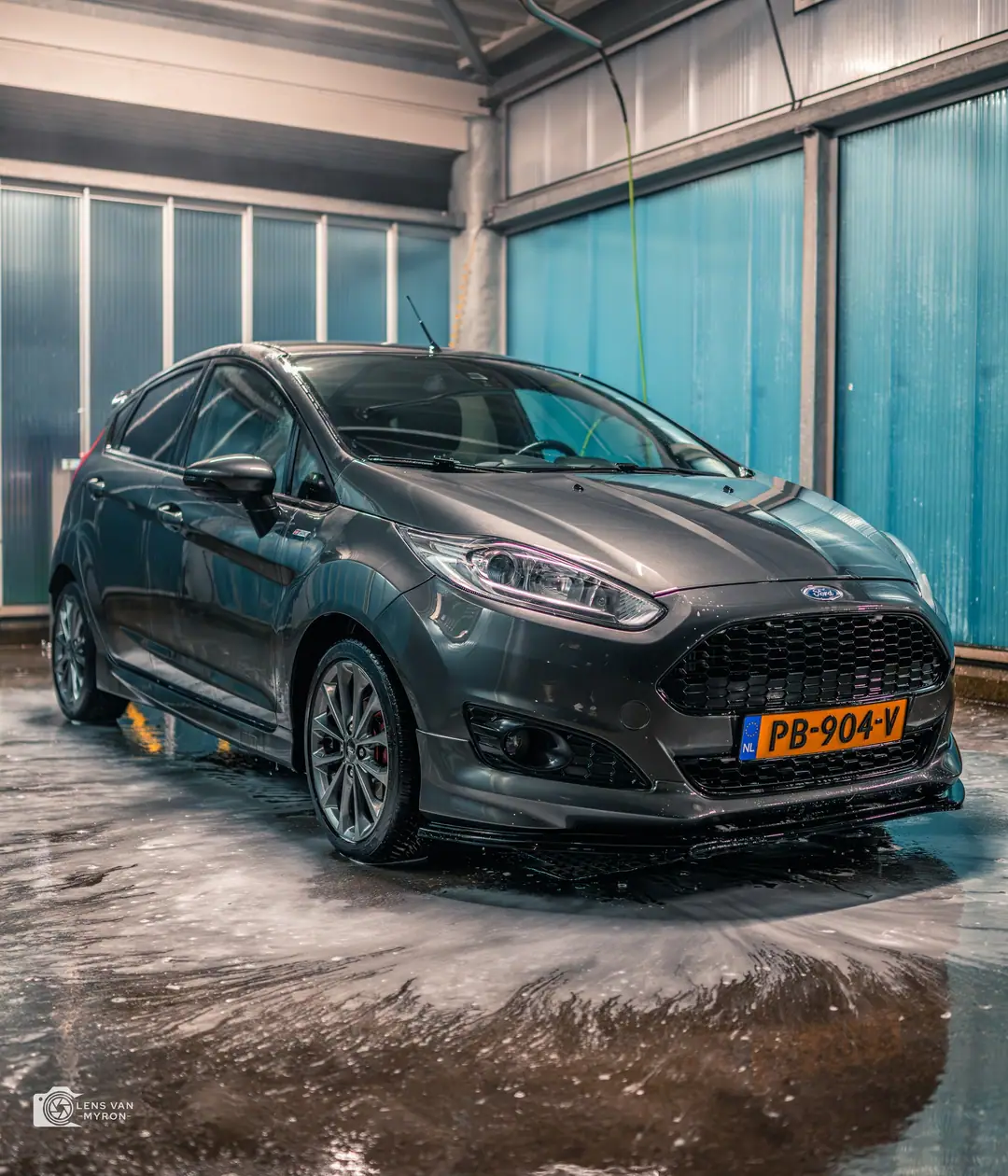 Donkergrijze Ford Fiesta in een wasbox met natte vloer, schuimresten en blauwe lichtdoorlatende wanden