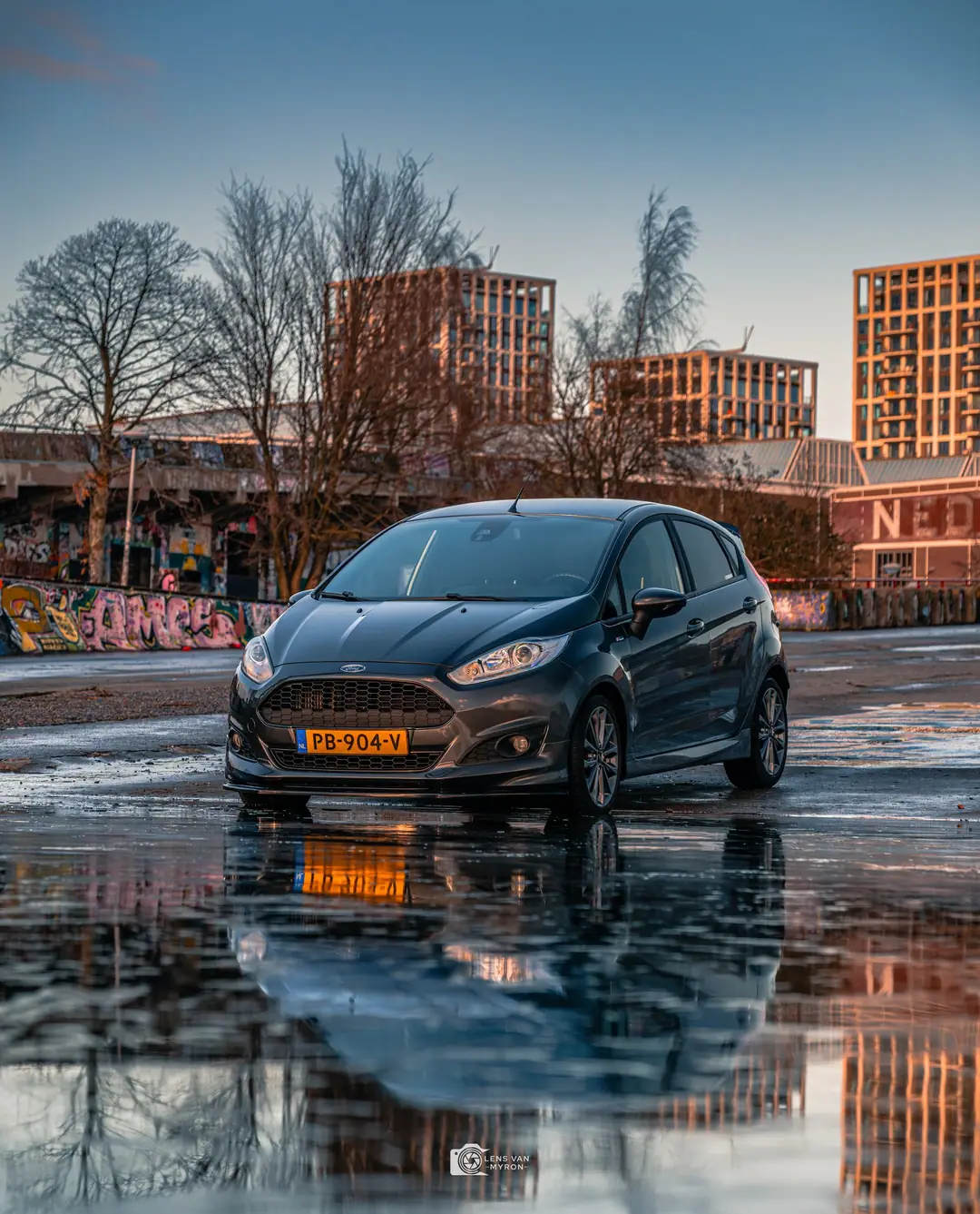 Donkergrijze Ford Fiesta op een nat plein met spiegeling in een plas, kale bomen, graffiti en hoge gebouwen in het laatste zonlicht