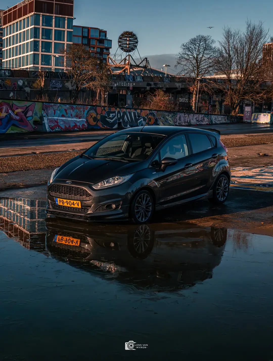 Ford Fiesta in laag winterlicht op een industrieterrein, weerspiegeld in een grote plas, met graffitimuur en moderne gebouwen erachter