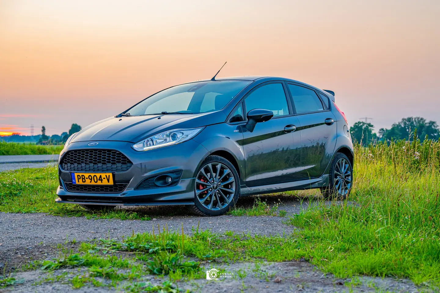 Donkergrijze Ford Fiesta op een grindpad tussen groen gras onder een pastelkleurige zonsondergang