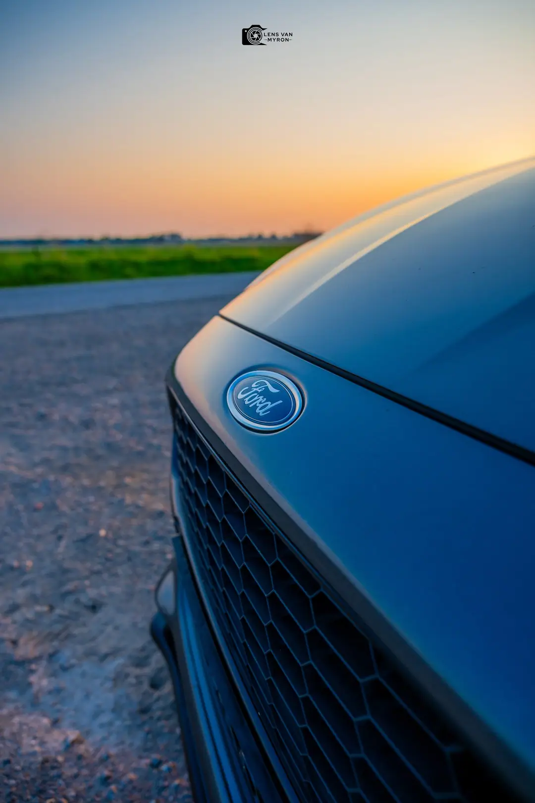 Close-up van het Ford-embleem en de grille met een onscherpe zonsondergang en weilanden op de achtergrond