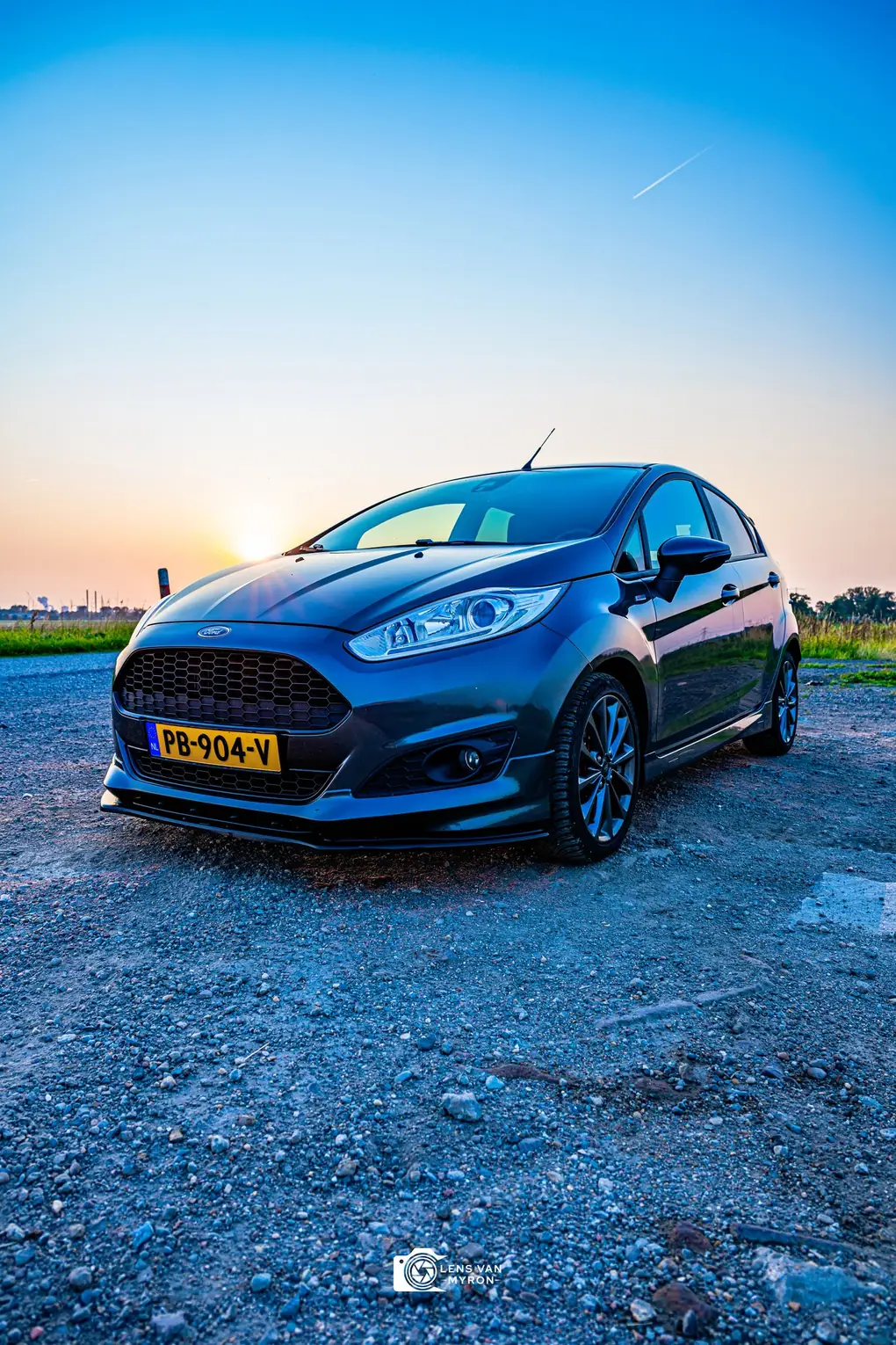 Ford Fiesta vanuit een lage hoek op grind, met een heldere blauwe lucht die naar oranje verloopt bij de horizon