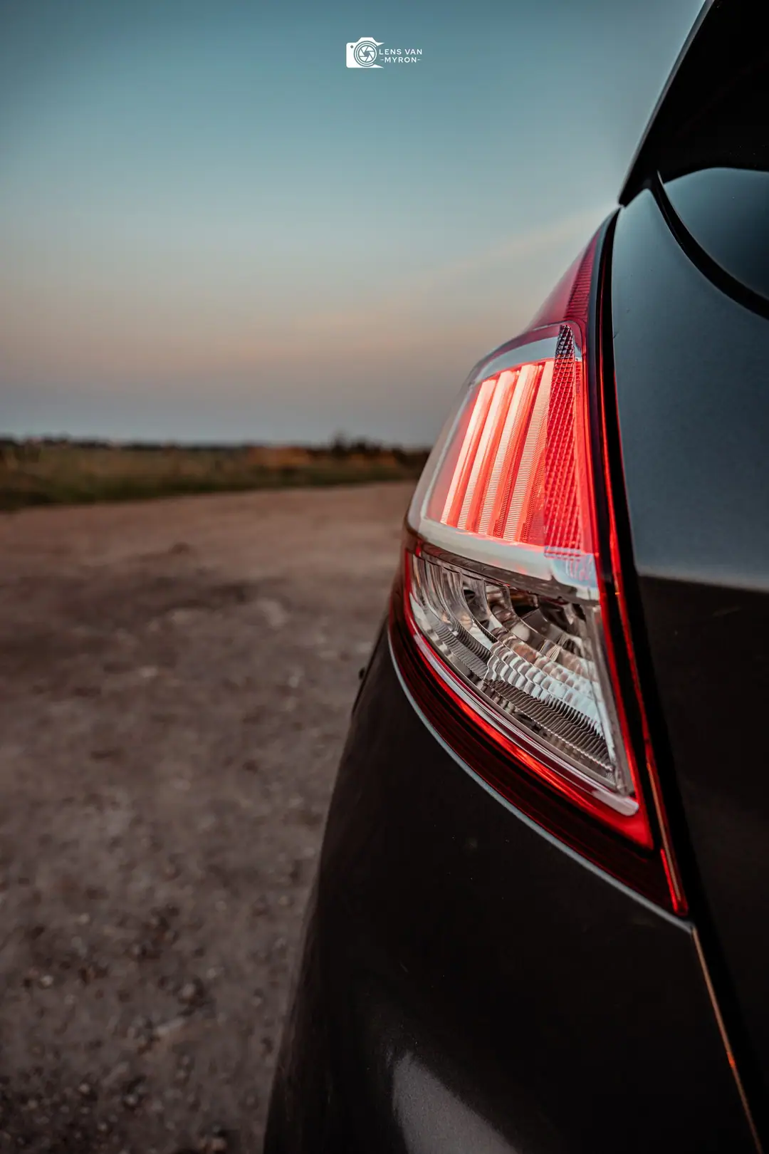 Detail van een rood achterlicht van een donkergrijze Ford Fiesta in de schemering, met een zandpad op de achtergrond