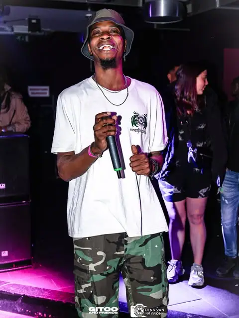 Een artiest met bucket hat en microfoon lacht op het podium van een club.