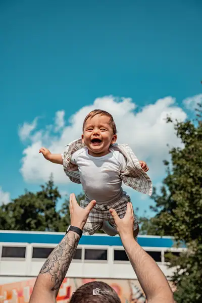 Levi wordt lachend hoog de lucht in gegooid door getatoeëerde armen, tegen een blauwe lucht met witte wolken.