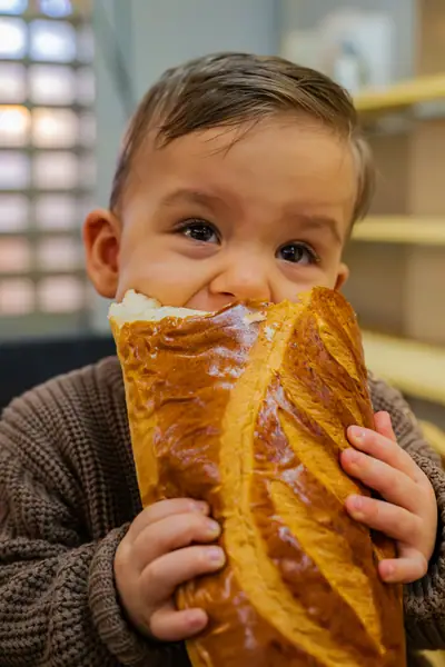 Levi houdt een groot stokbrood vast en kijkt er met grote ogen bovenuit, in een bruine gebreide trui.