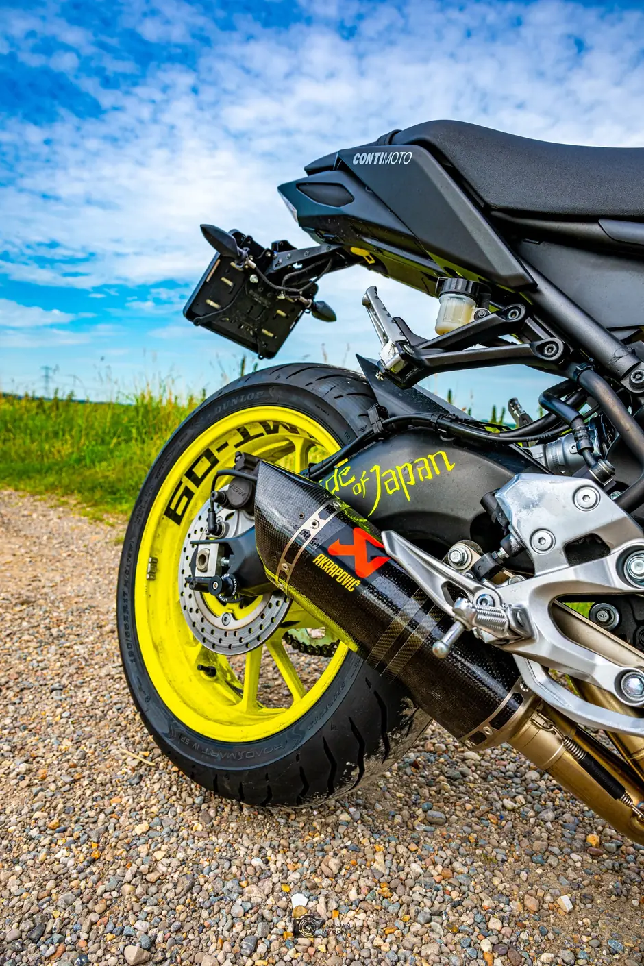 Detail van het geel gespoten achterwiel en de carbon Akrapovič-uitlaat van een Yamaha MT-09 op een grindpad, onder een blauwe lucht met wolken.