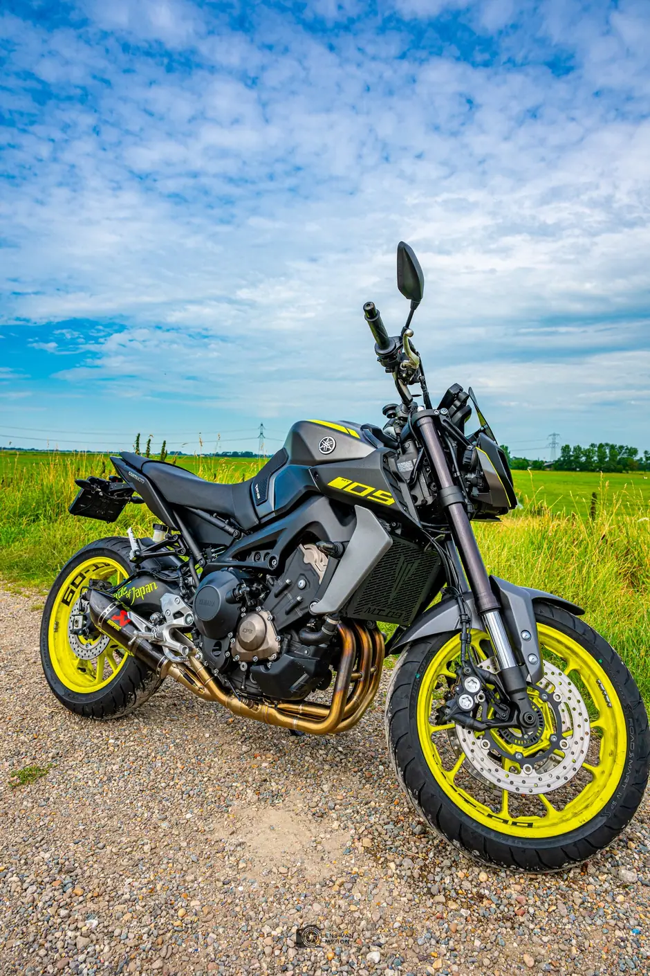 Zwarte Yamaha MT-09 met gele velgen en goudkleurige uitlaatbochten op een grindpad tussen het gras, onder een wijde blauwe lucht met wolken.