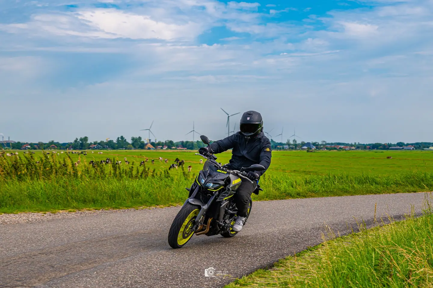 Motorrijder met zwarte helm en jas neemt op een Yamaha MT-09 een bocht op een smal polderweggetje, met koeien en windmolens op de achtergrond.