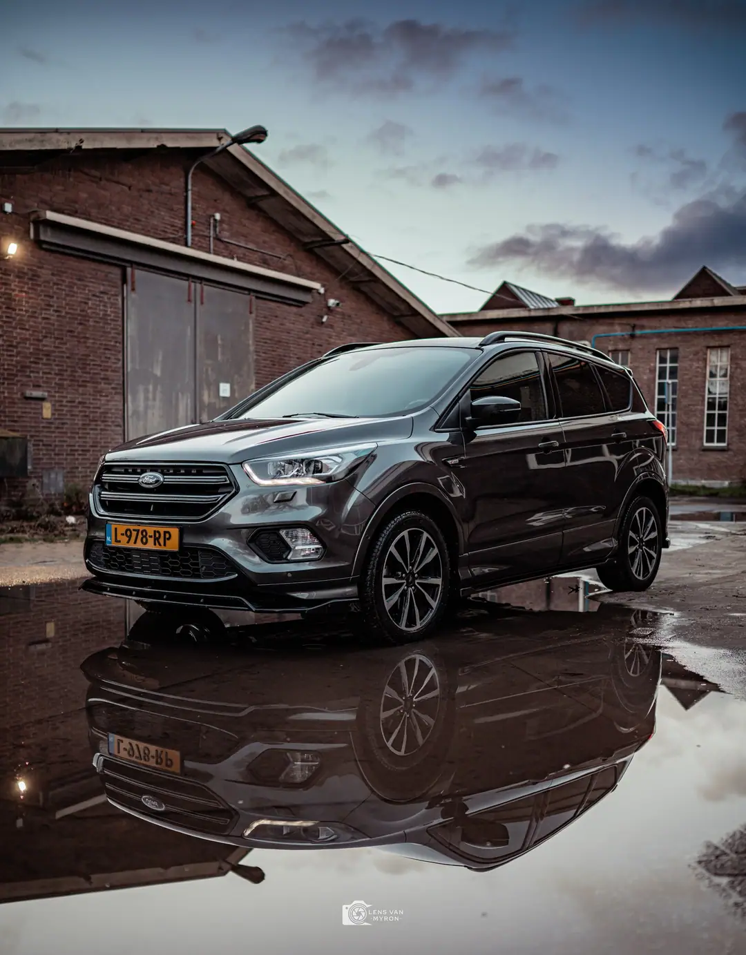 Grijze Ford Kuga voor oude bakstenen fabriekshallen, volledig weerspiegeld in een grote plas onder een bewolkte avondlucht