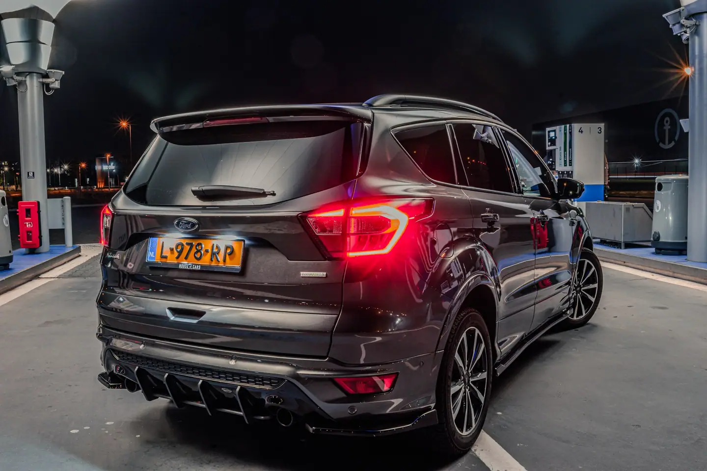Ford Kuga van achteren bij nacht op een tank- of laadstation, met fel oplichtende achterlichten en een straatlantaarn met lichtster