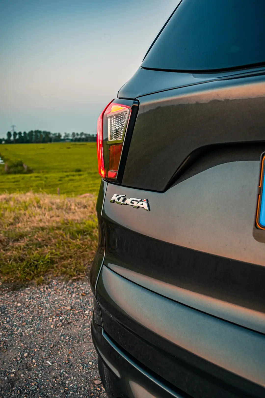 Detail van de achterkant van een Ford Kuga met het chromen Kuga-logo en het achterlicht, met groene weilanden op de achtergrond