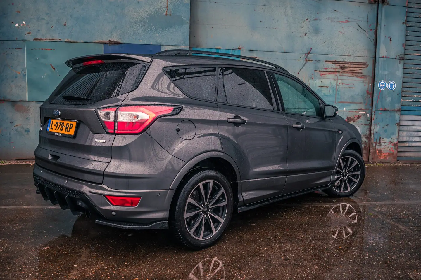 Grijze Ford Kuga schuin van achteren voor een verweerde blauwe bedrijfsmuur op nat asfalt