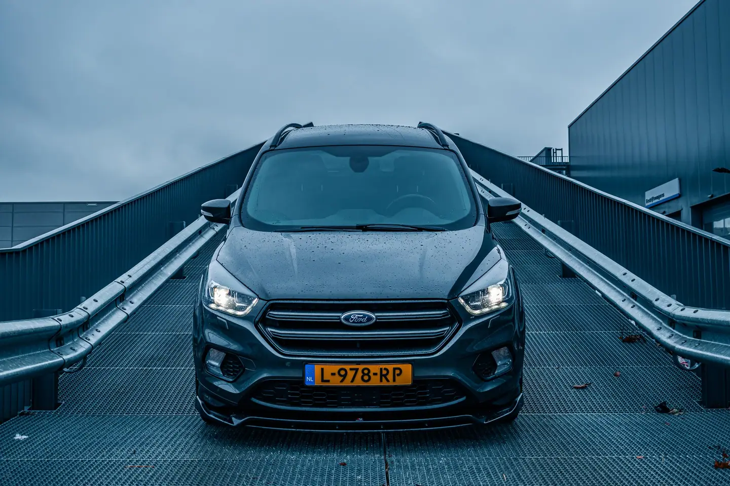 Ford Kuga recht van voren op een stalen hellingbaan tussen vangrails, met brandende koplampen onder een grijze lucht