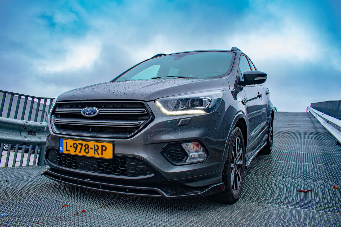 Ford Kuga vanuit een lage hoek op een stalen hellingbaan onder een dramatische blauwe wolkenlucht