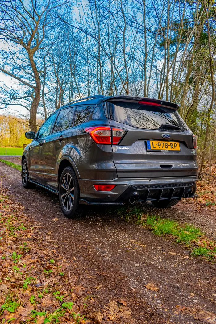 Ford Kuga schuin van achteren op een bospad met afgevallen bladeren, kale bomen en een blauwe lucht