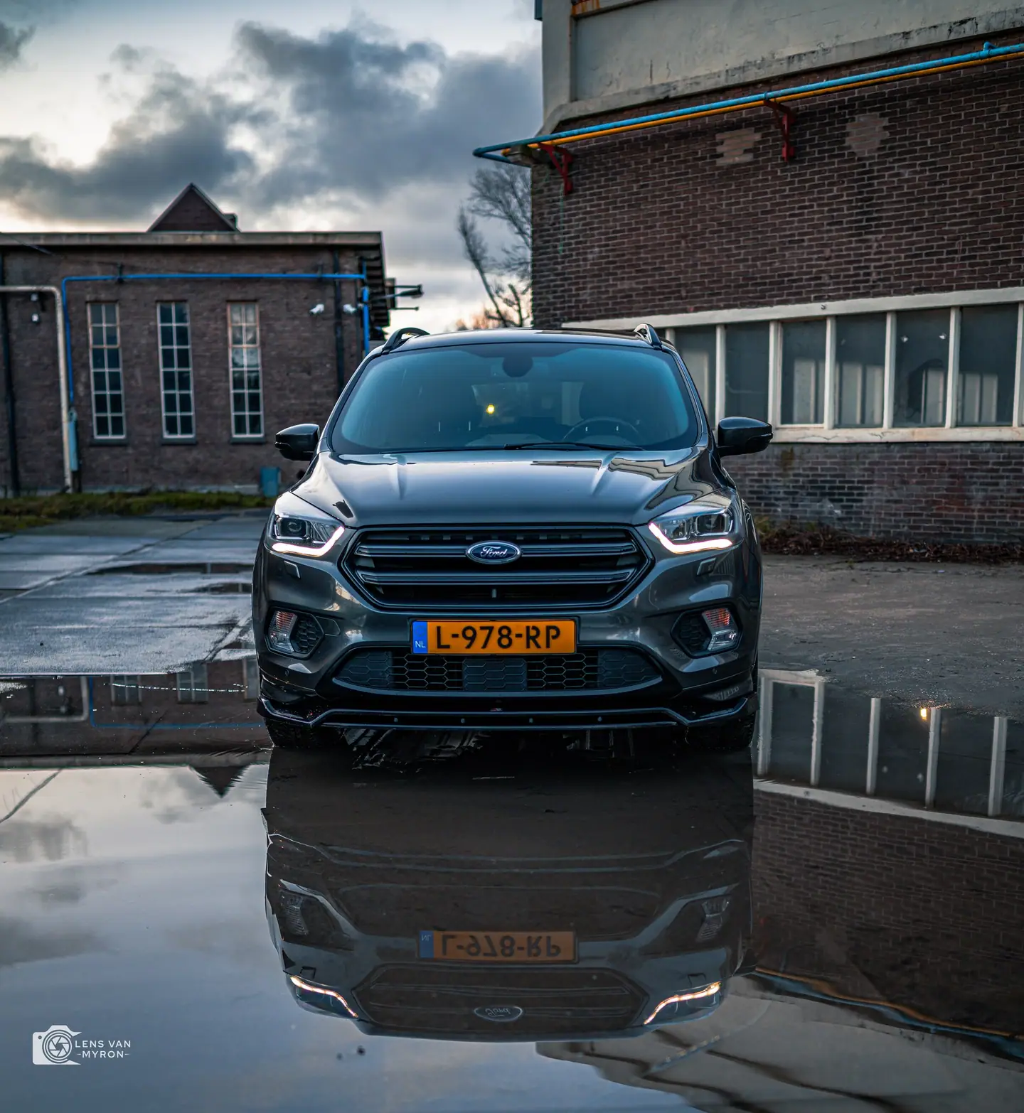 Ford Kuga recht van voren tussen bakstenen fabrieksgebouwen, perfect gespiegeld in een plas onder een dramatische avondlucht
