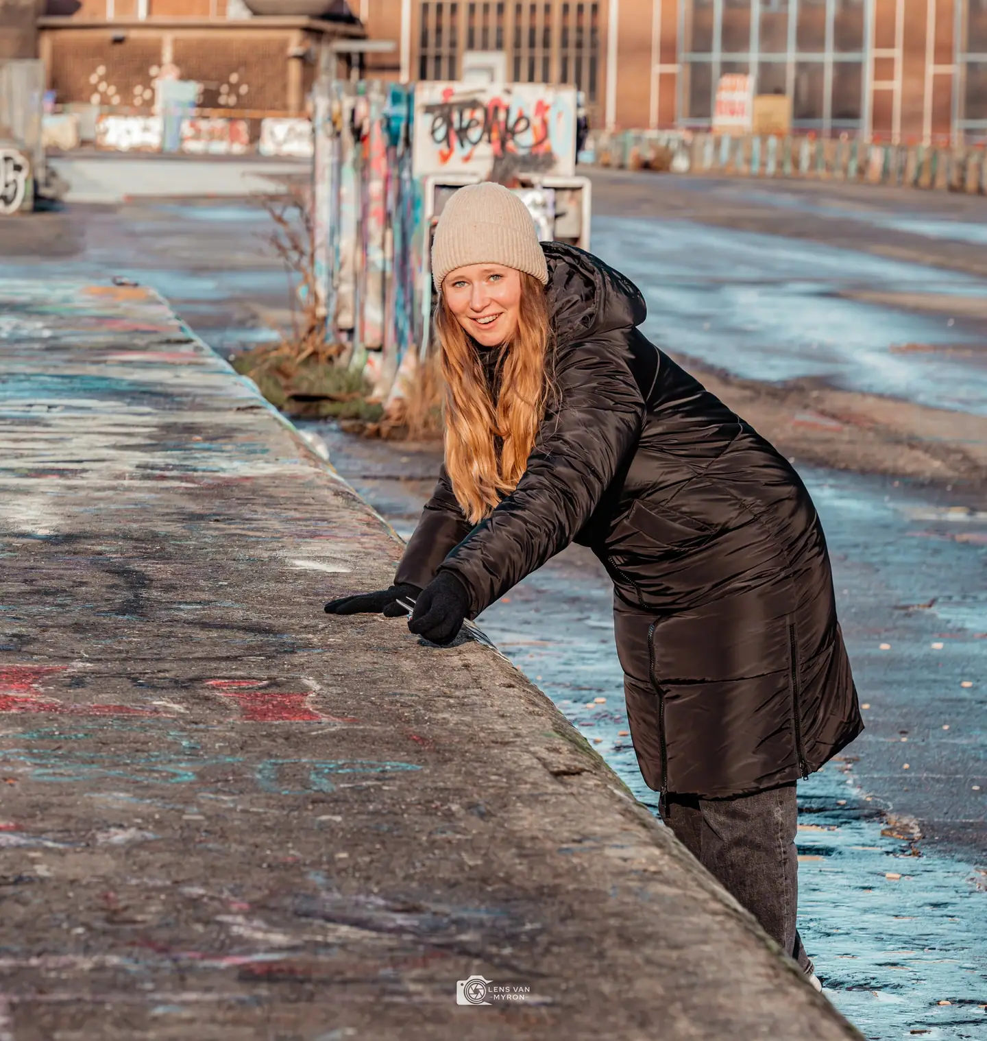 Nienke met beige muts en lange zwarte winterjas leunt lachend over een met graffiti bespoten betonnen rand op een industrieel terrein.