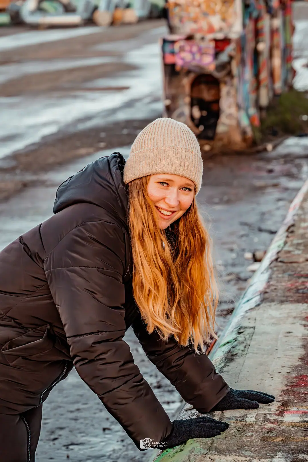 Nienke leunt met handschoenen op een betonnen rand en lacht naar de camera, haar lange roodblonde haar verlicht door de laagstaande zon.