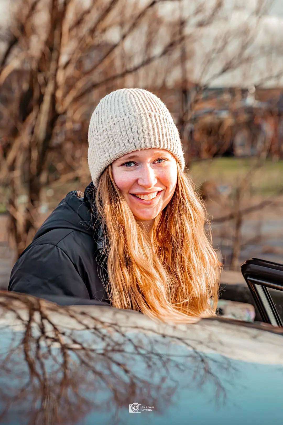 Nienke kijkt lachend over het dak van een auto waarin kale bomen weerspiegelen.