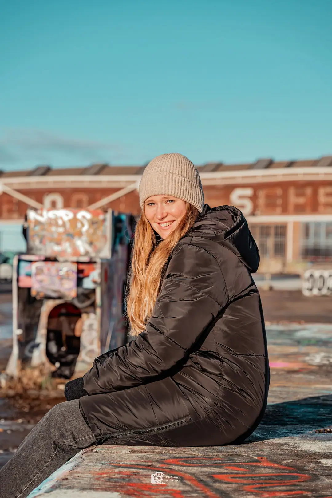 Nienke zit op een betonnen rand en kijkt lachend over haar schouder, met graffiti, een industriële hal en een blauwe lucht achter haar.