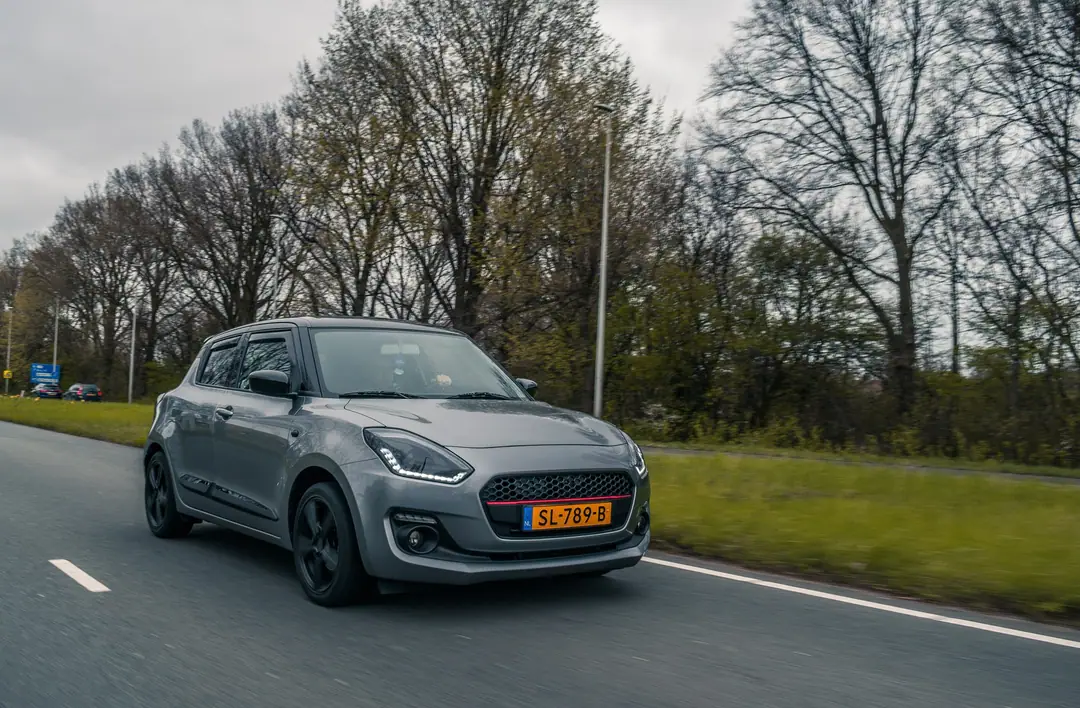 Grijze Suzuki Swift met zwarte velgen rijdend op een weg langs bomen, met bewegingsonscherpte in het wegdek.