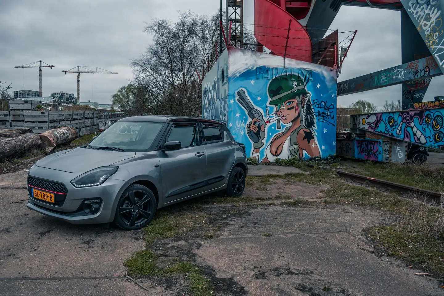 Grijze Suzuki Swift laag van voren gefotografeerd op een ruig industrieel terrein naast een grote graffitimuurschildering en bouwkranen.
