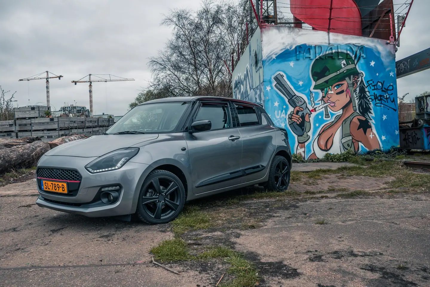 Grijze Suzuki Swift met zwarte velgen schuin van voren voor een kleurrijke muurschildering onder een bewolkte lucht.