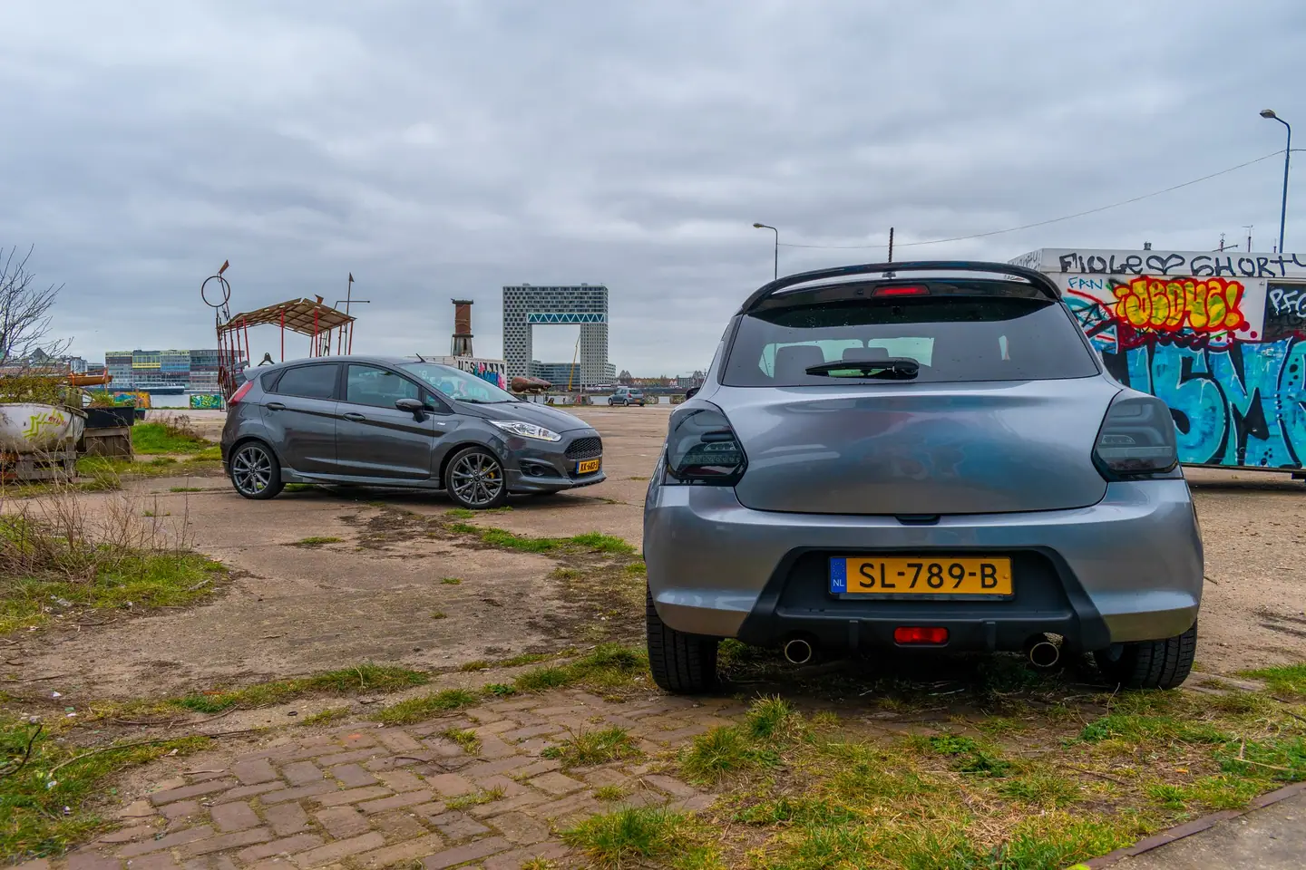 Achterkant van een grijze Suzuki Swift op een industrieel terrein aan het water, met een donkergrijze Ford Fiesta en moderne gebouwen op de achtergrond.
