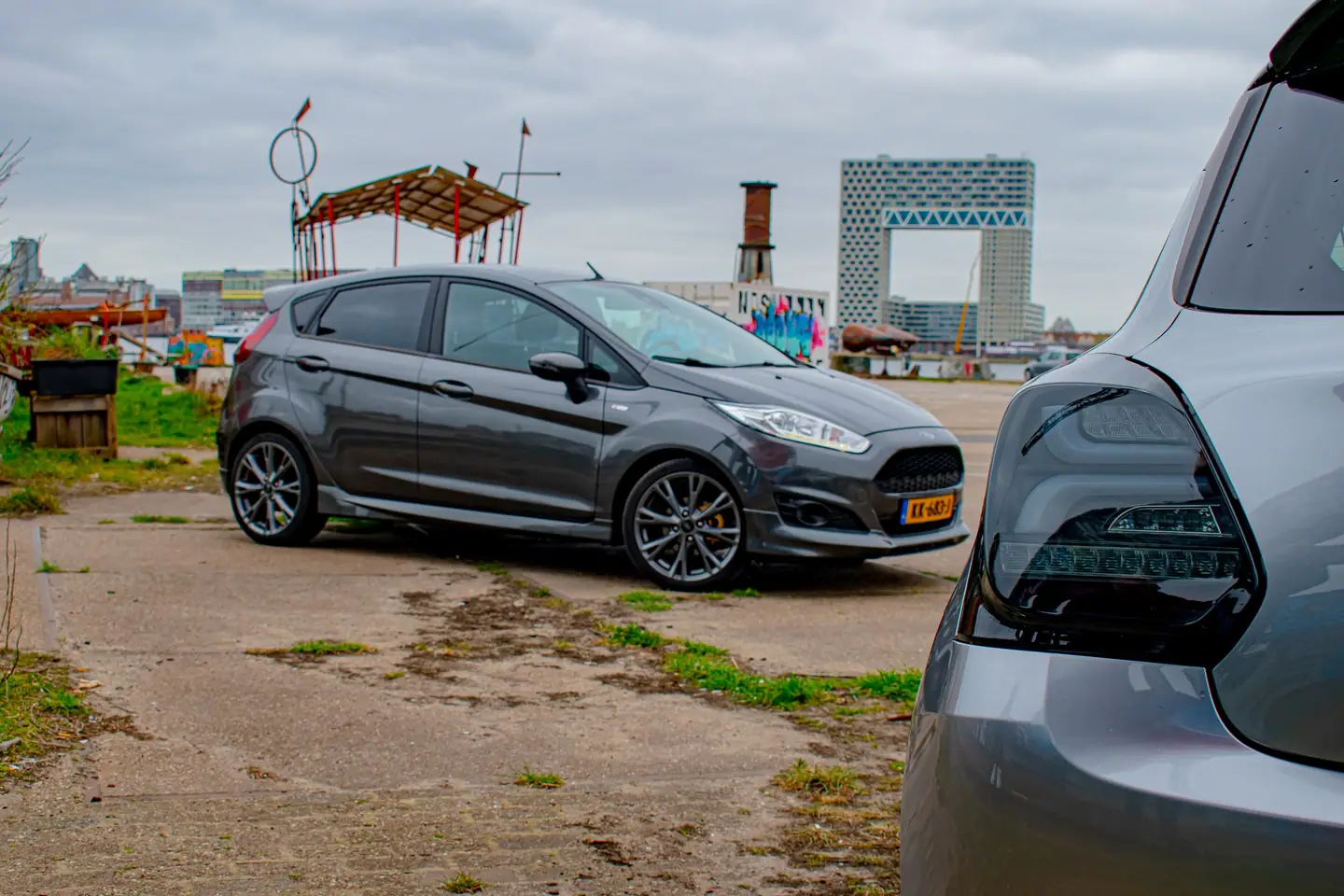 Donkergrijze Ford Fiesta op een verlaten kade, met op de voorgrond het onscherpe achterlicht van een grijze Suzuki Swift.