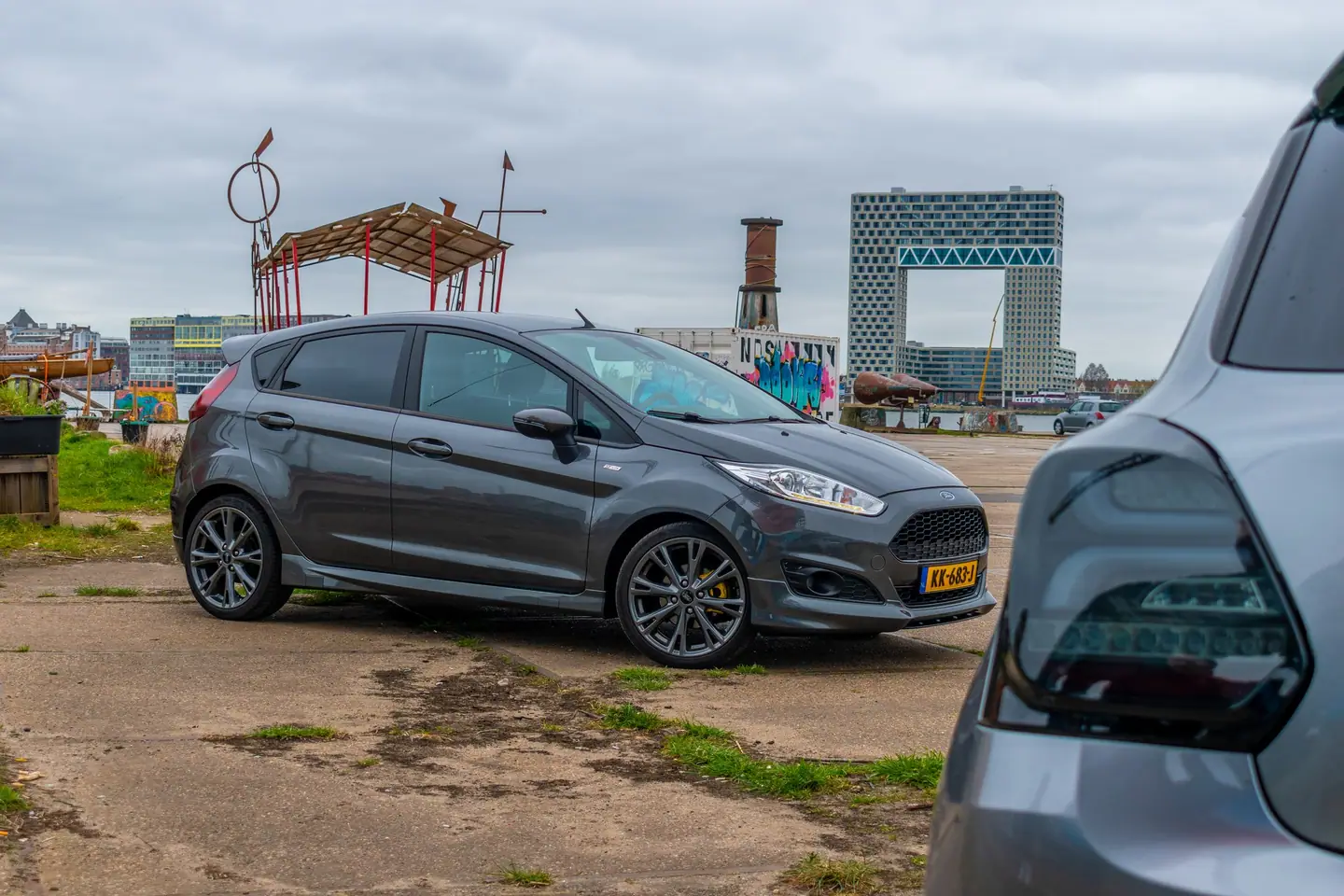 Donkergrijze Ford Fiesta met lichtmetalen velgen en gele remklauwen op een kade, met rechts het achterlicht van een grijze Suzuki Swift.