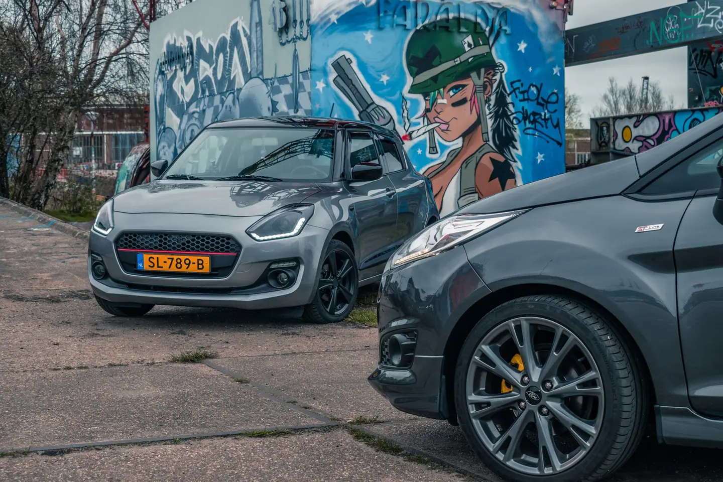 Grijze Suzuki Swift voor een graffitimuurschildering, met op de voorgrond het voorwiel en de flank van een donkergrijze Ford Fiesta.