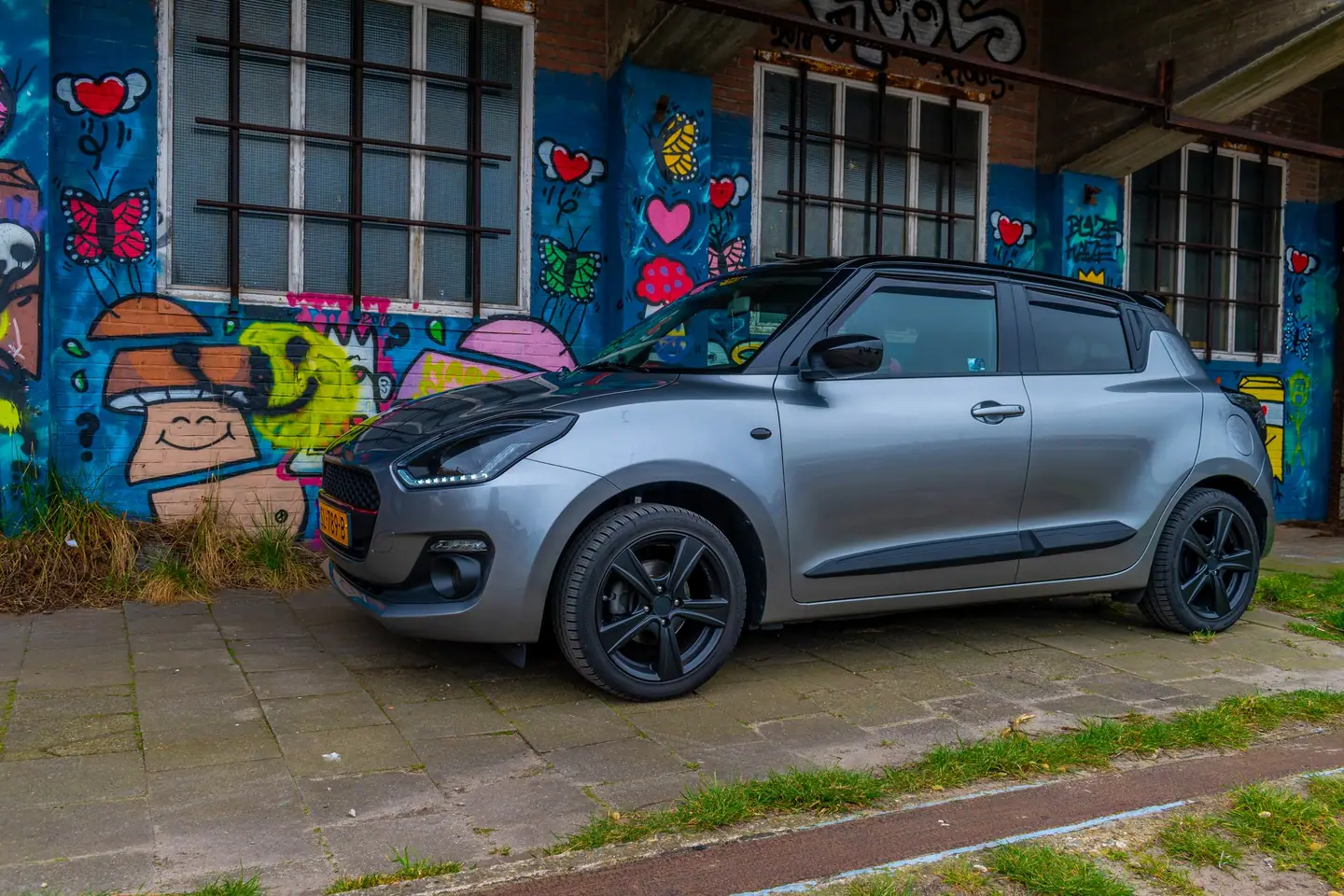Grijze Suzuki Swift met zwarte velgen in zijaanzicht voor een muur met kleurrijke graffiti van harten, vlinders en paddenstoelen.