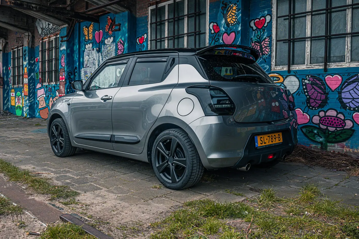 Grijze Suzuki Swift schuin van achteren met dakspoiler en zwarte velgen, voor een muur vol kleurrijke graffiti met harten en bloemen.
