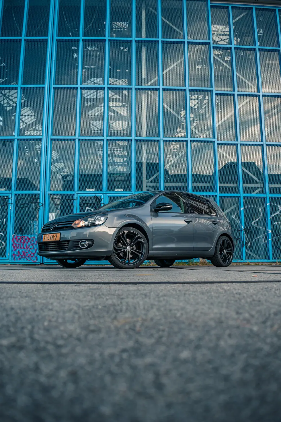 Grijze Volkswagen Golf 6 met zwarte velgen, laag van voren gefotografeerd voor een gevel van blauwe stalen kozijnen en glas.