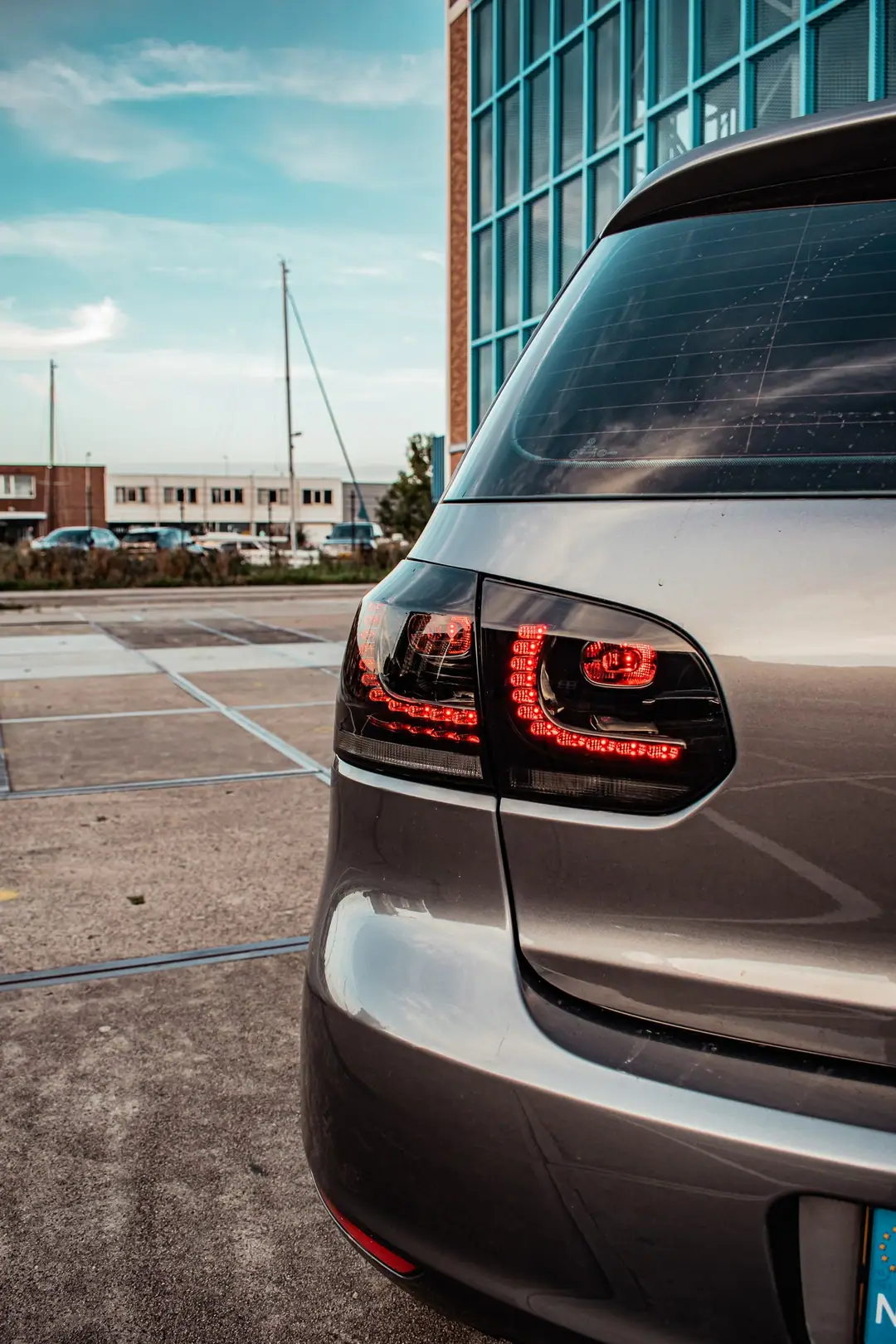 Close-up van de rode LED-achterlichten van een grijze Volkswagen Golf 6, met op de achtergrond een parkeerterrein en scheepsmasten.