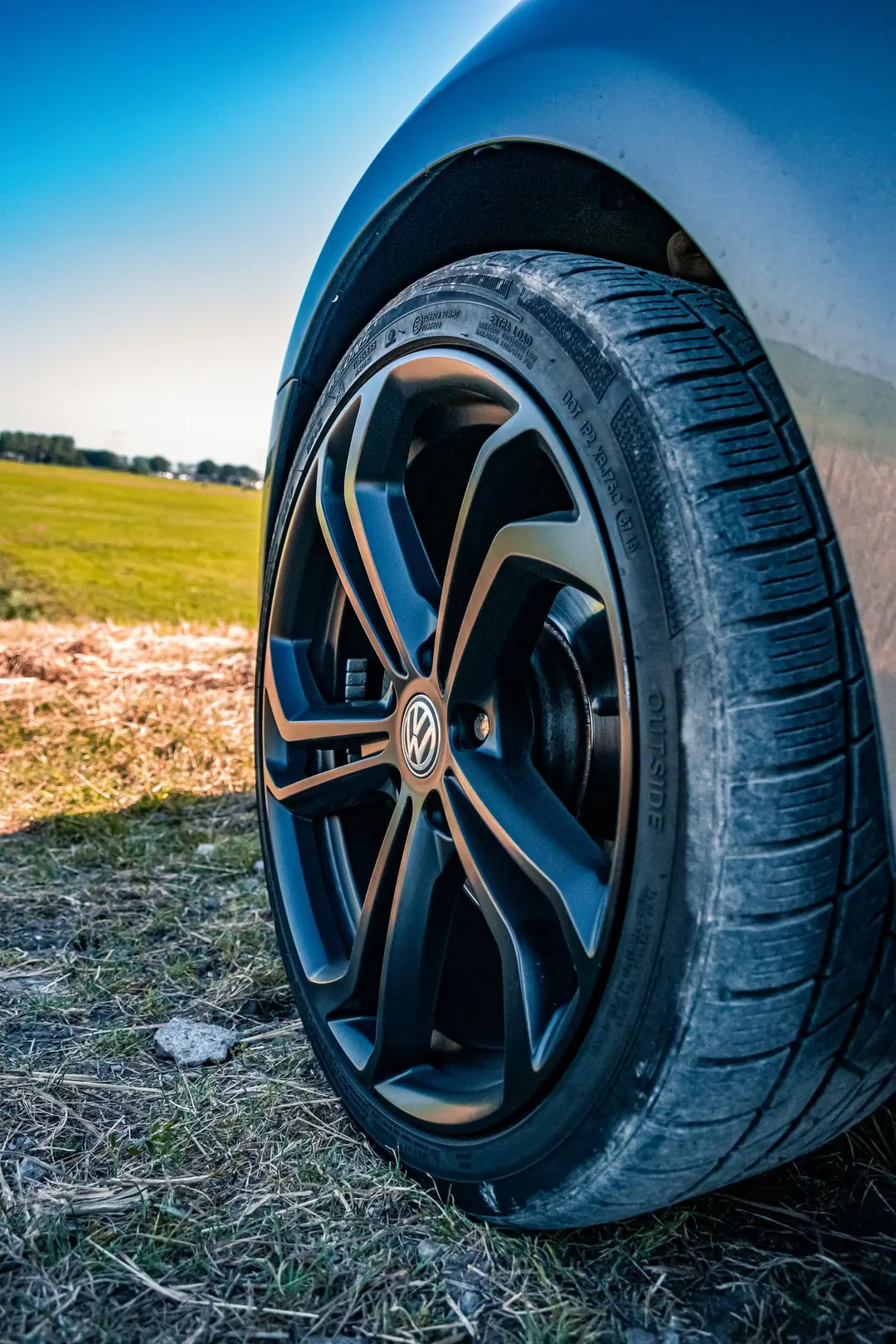 Zwarte Volkswagen-lichtmetalen velg met band van dichtbij, in het gras met een weiland en blauwe lucht op de achtergrond.