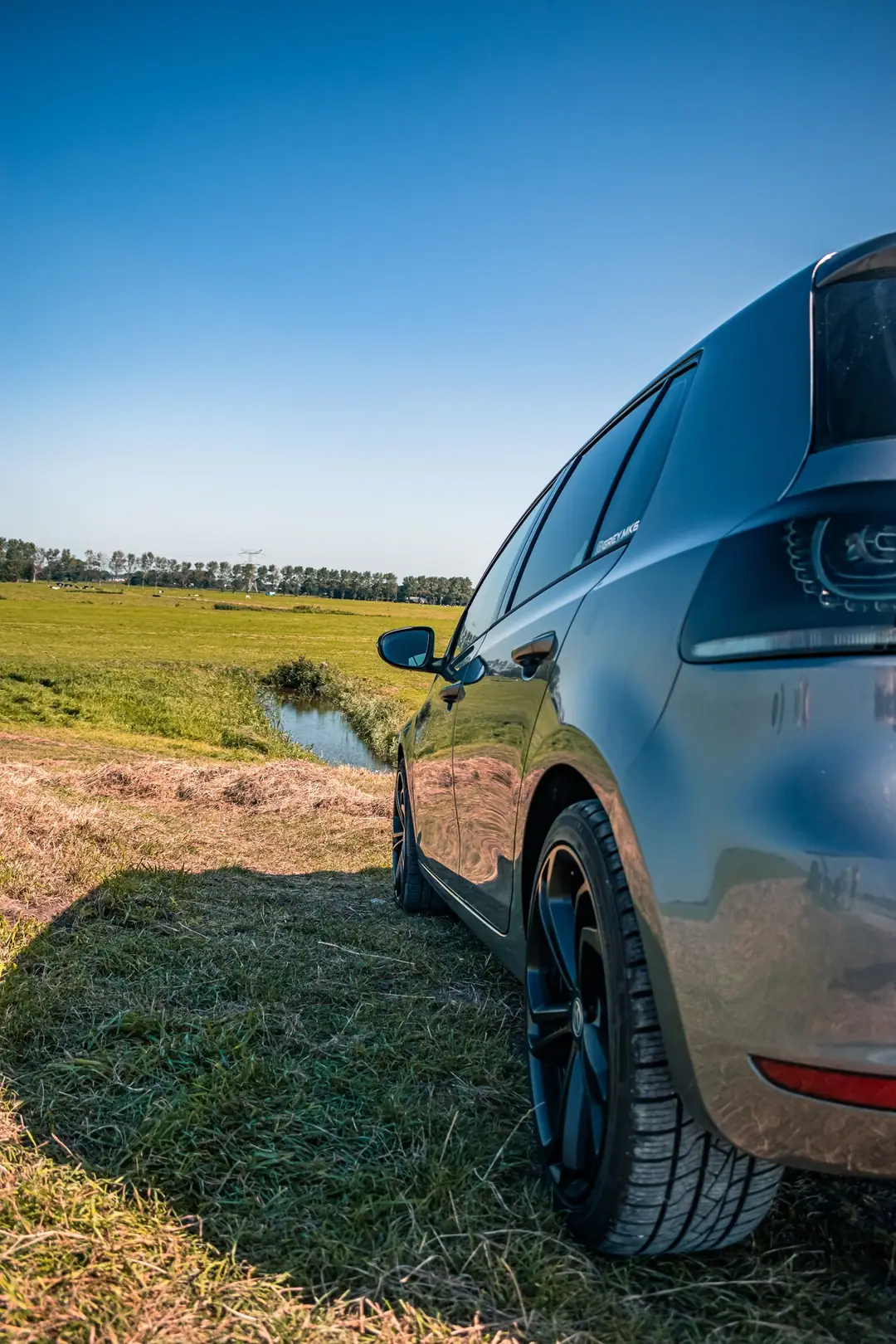 Achterkant van een grijze Volkswagen Golf 6 op het gras naast een sloot in een vlak Hollands weiland onder een strakblauwe lucht.