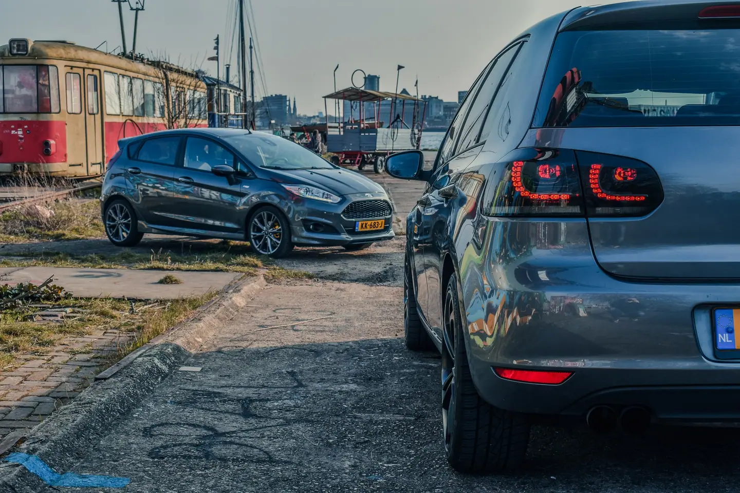 Achterlichten van een grijze Volkswagen Golf 6 op de voorgrond, met een grijze Ford Fiesta, een oude tram en de stadsskyline aan het water op de achtergrond.