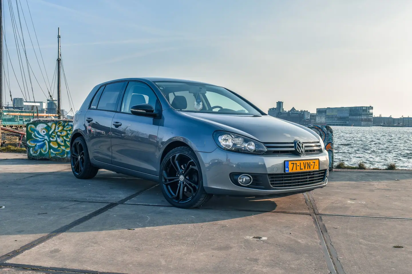 Grijze Volkswagen Golf 6 met zwarte velgen schuin van voren op een kade, met water en moderne gebouwen in zacht, laag zonlicht.