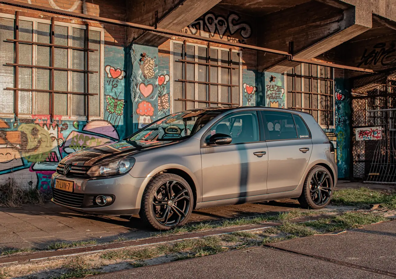 Grijze Volkswagen Golf 6 in warm zonlicht voor een muur vol kleurrijke graffiti met harten, vlinders en paddenstoelen.