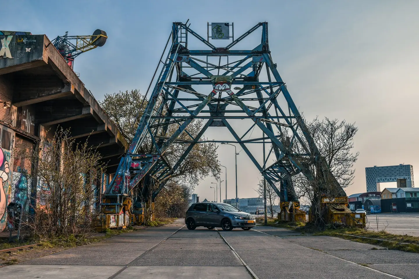 Grijze Volkswagen Golf 6 klein in beeld onder een grote stalen kraanconstructie op een industrieel terrein met graffiti, onder een zachte pastellucht.
