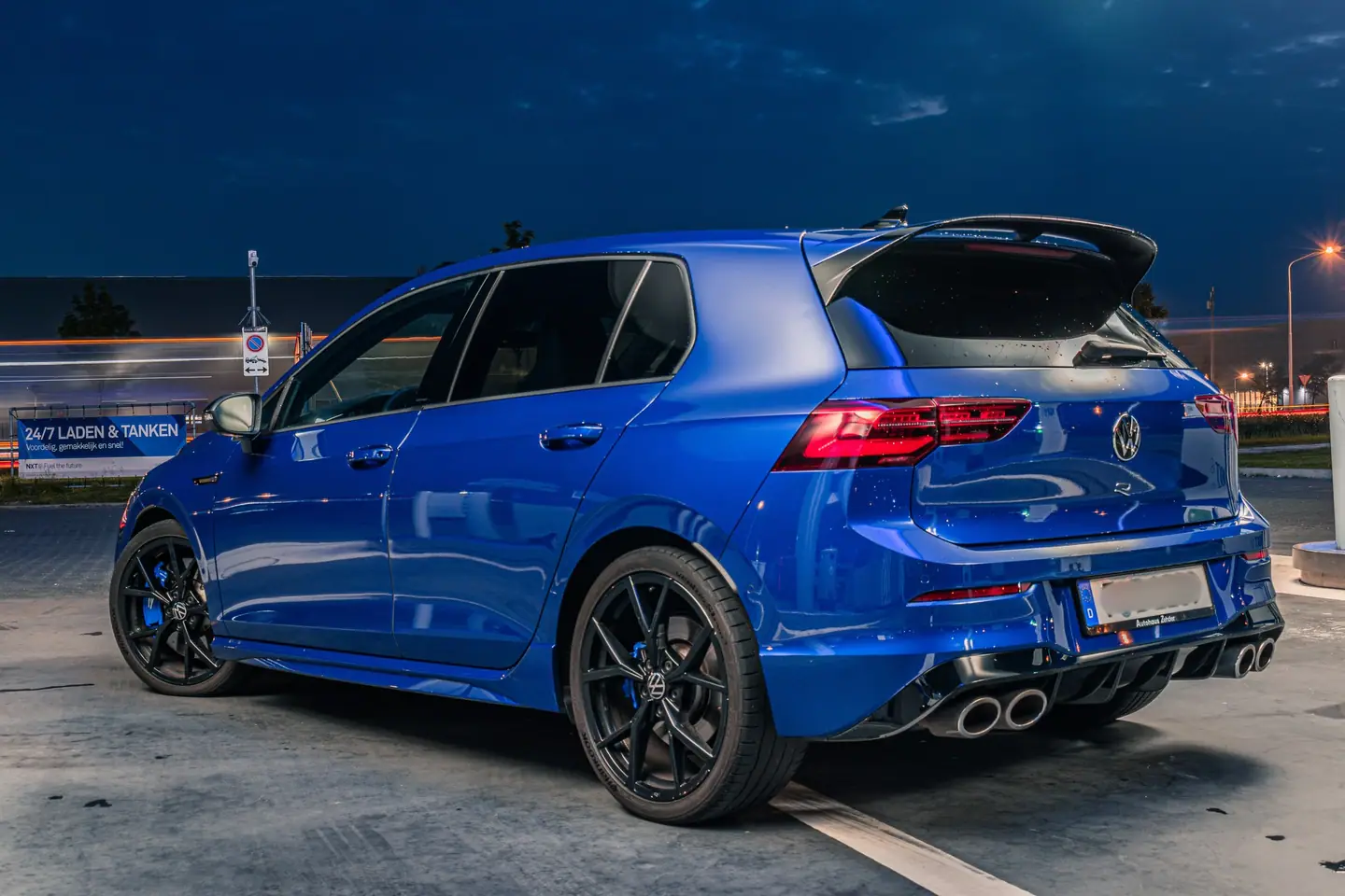 Blauwe Volkswagen Golf R schuin van achteren tijdens het blauwe uur bij een tankstation, met lichtsporen van passerend verkeer op de achtergrond