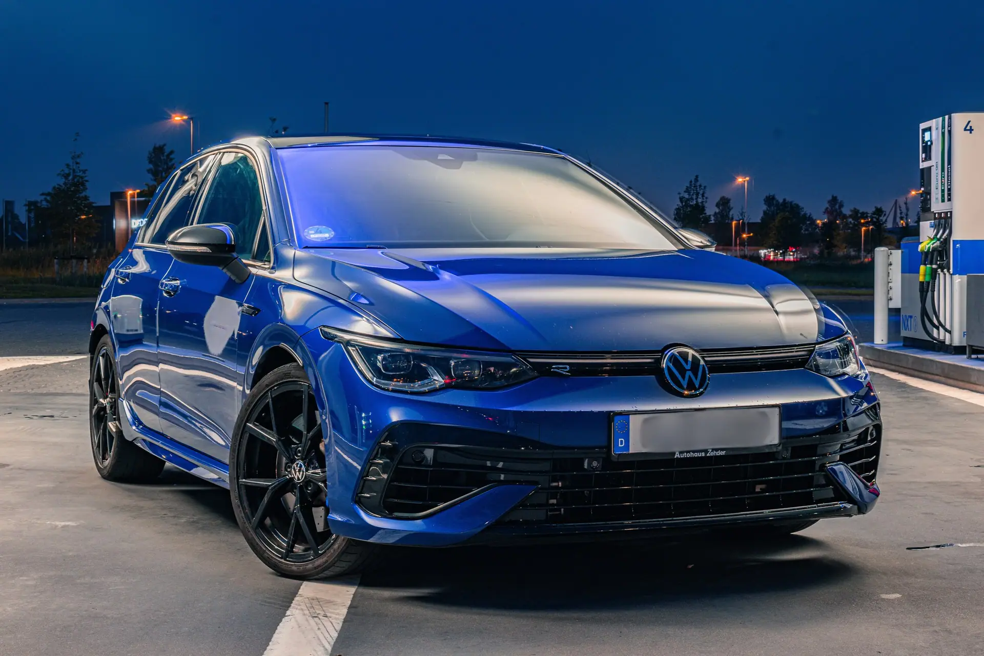 Blauwe Volkswagen Golf R schuin van voren tijdens het blauwe uur op het terrein van een tankstation